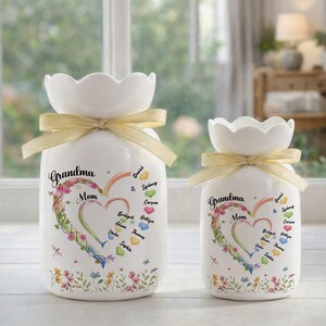 May include: Two white ceramic vases with a scalloped top and a yellow ribbon tied around the neck. The vases are decorated with a heart-shaped floral design and the words "Grandma Mom". Names are written inside and around the heart.