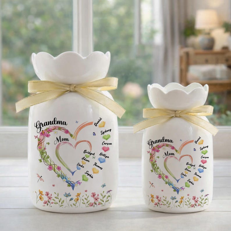 May include: Two white ceramic vases with a scalloped top and a yellow ribbon tied around the neck. The vases are decorated with a heart-shaped floral design and the words "Grandma Mom". Names are written inside and around the heart.