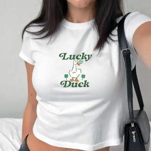 May include: White cropped t-shirt with a green graphic of a duck holding a four-leaf clover and the text "Lucky Duck".