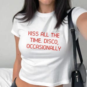 May include: White crop top with the red text "KISS ALL THE TIME. DISCO, OCCASIONALLY." The shirt has short sleeves and a round neckline. A black shoulder bag is visible on the right side of the image.