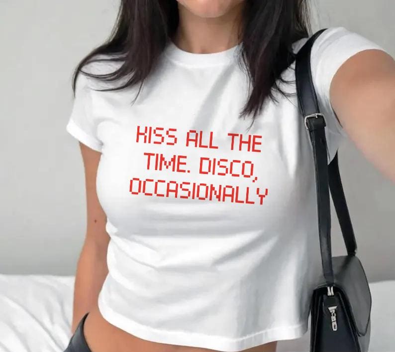 May include: White crop top with the red text "KISS ALL THE TIME. DISCO, OCCASIONALLY." The shirt has short sleeves and a round neckline. A black shoulder bag is visible on the right side of the image.