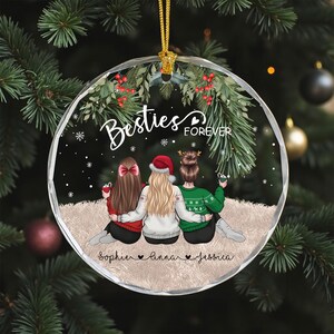 Pu&ograve; includere: Una palla di Natale rotonda e trasparente con un filo d'oro. L'ornamento presenta tre figure con il testo "Besties Forever" e i nomi "Sophie, Linna, Jessica". Le figure indossano maglioni e un cappello da Babbo Natale.