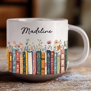 May include: White ceramic mug with a brown base and a handle. The mug features a colorful design of books and flowers, with the name "Madeline" written in cursive. The design wraps around the mug, creating a charming and personalized look.