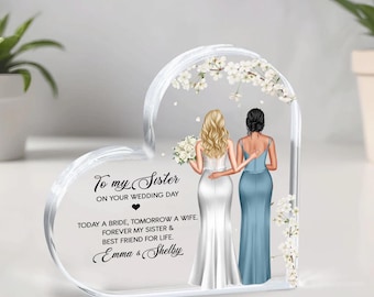May include: A heart-shaped clear acrylic keepsake with a white floral design and two women in wedding attire. The text reads "To my Sister on your Wedding Day. Today a bride, tomorrow a wife. Forever my sister & best friend for life. Emma & Shelby."