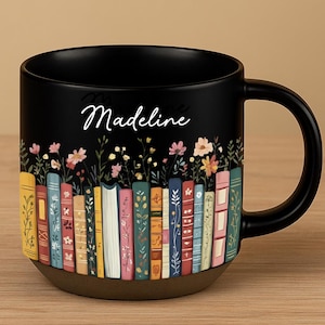 May include: Black ceramic mug featuring a colorful floral and book design. The mug has a brown base and a black handle. The name "Madeline" is written in cursive on the mug.