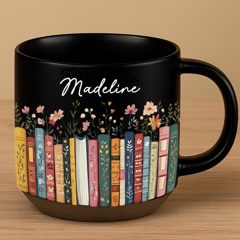 May include: Black ceramic mug featuring a colorful floral and book design. The mug has a brown base and a black handle. The name "Madeline" is written in cursive on the mug.