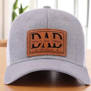 May include: Grey trucker cap with a white mesh back. A brown leather patch on the front displays the word "DAD" and the names Robin, Emily, and Taylor. The text "We love you" is also on the patch. A classic baseball cap style.