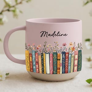 May include: A pink ceramic mug with a light beige base. The mug features a colorful illustration of books and flowers. The name "Madeline" is written in black script on the side of the mug.