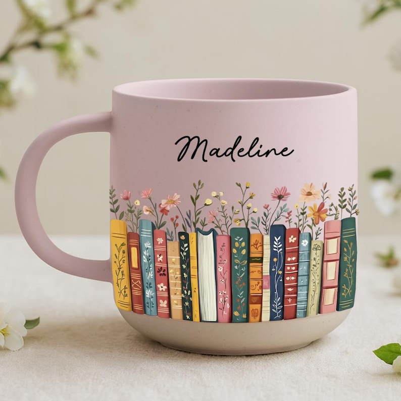 May include: A pink ceramic mug with a light beige base. The mug features a colorful illustration of books and flowers. The name "Madeline" is written in black script on the side of the mug.