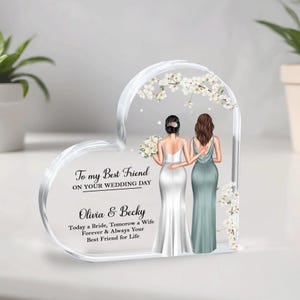 May include: A heart-shaped clear acrylic keepsake with a white and green illustration of two women, one in a white wedding dress and the other in a green bridesmaid dress. The text reads "To my Best Friend on your Wedding Day Olivia & Becky Today a Bride, Tomorrow a Wife Forever & Always Your Best Friend for Life."
