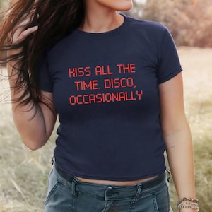 May include: Navy blue t-shirt with the text "KISS ALL THE TIME. DISCO, OCCASIONALLY" in red pixelated font. The shirt is a crew neck style and is made of a soft material. The model is wearing blue jeans.