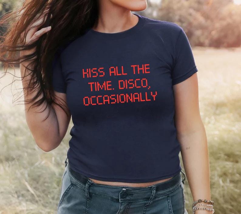 May include: Navy blue t-shirt with the text "KISS ALL THE TIME. DISCO, OCCASIONALLY" in red pixelated font. The shirt is a crew neck style and is made of a soft material. The model is wearing blue jeans.