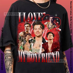 Op de afbeelding: Een zwart T-shirt met een grafische print van een groep mannen met de tekst "I Love My Boyfriend" in rood en wit. De grafiek is omringd door rode harten en bliksem.