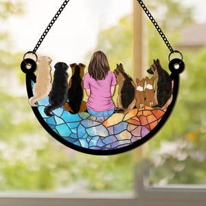 Puede incluir: Un adorno colgante decorativo que representa a una mujer y varios perros. Los perros son de diferentes razas y colores, sentados juntos. El adorno tiene un borde negro y una cadena para colgar. El fondo es un diseño de vidriera.