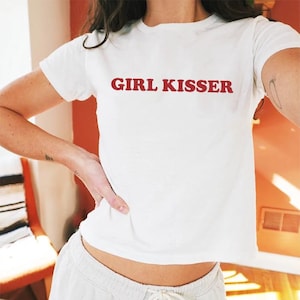 Puede incluir: Camiseta blanca con el texto "GIRL KISSER" en letras rojas. La camiseta es de manga corta y parece estar hecha de un material suave. La persona lleva pantalones de color claro.