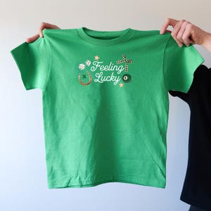 Può includere: Maglietta verde con una grafica bianca che dice "Feeling Lucky" e presenta un ferro di cavallo, dadi, un mulino a vento e una palla da biliardo.