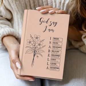 Puede incluir: Un diario de color melocotón con las palabras "God say I am" en escritura negra. Una ilustración floral y la palabra "ISABELLA" están debajo del texto. El diario enumera las palabras "Image, Saved, Abundant, Beloved, Equipped, Light, Loved, Accepted".