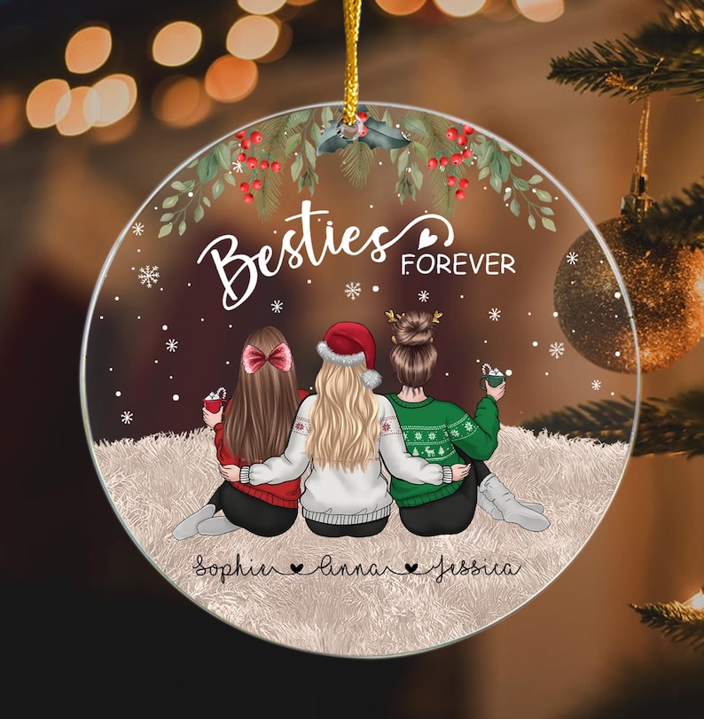 Pu&ograve; includere: Un ornamento natalizio rotondo con le parole "Besties Forever" e i nomi Sophie, Anna e Jessica. L'ornamento presenta tre figure in abiti invernali, con un design festivo.