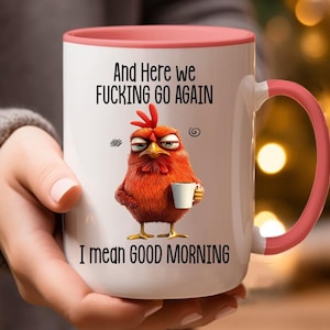 May include: White ceramic coffee mug with a pink rim and handle. The mug features a cartoon rooster with a grumpy expression holding a cup of coffee. The text on the mug reads "And here we fucking go again I mean good morning."