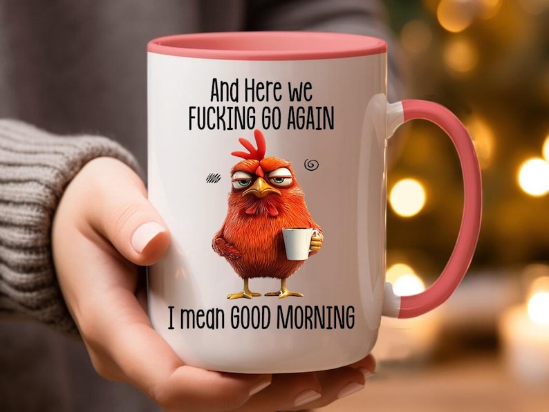 May include: White ceramic coffee mug with a pink rim and handle. The mug features a cartoon rooster with a grumpy expression holding a cup of coffee. The text on the mug reads "And here we fucking go again I mean good morning."