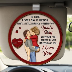 May include: A round car visor ornament with a red border and a wooden appearance. The ornament features a couple embracing, with text that reads "You're Sexy, I Love You." A heart-shaped puzzle piece with the names "William" and "Sophia" is also visible.