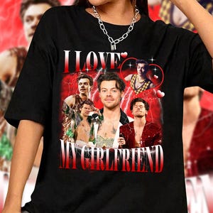 Op de afbeelding: Zwart T-shirt met een grafische print van een collage van afbeeldingen van een man met de tekst "I LOVE MY GIRLFRIEND" in rode en witte letters.