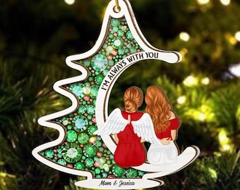 May include: A white wooden Christmas ornament shaped like a tree with green and red glitter. Two women with angel wings are sitting on a crescent moon with the text "I'm always with you" and "Mom & Jessica".