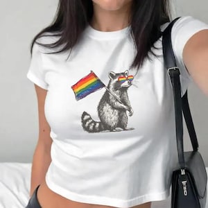 Może przedstawiać: Biały top z nadrukiem szopa pracza w tęczowych okularach przeciwsłonecznych i trzymającego tęczową flagę. Szop stoi. Koszulka ma okrągły dekolt.