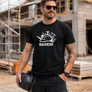 May include: A black short-sleeved t-shirt featuring a white graphic print of a construction helmet surrounded by various hand tools and the text "BAUHERR" centred below the helmet design. The shirt has a crew neckline and a standard fit.