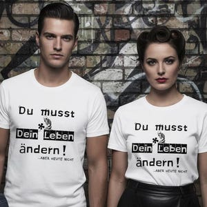 Könnte beinhalten: Zwei Personen tragen weiße T-Shirts mit dem deutschen Text "Du musst Dein Leben ändern!" in Schwarz. Die Shirts haben eine Grafik und den Text "Aber heute nicht" darunter. Sie stehen vor einer Backsteinmauer mit Graffiti.