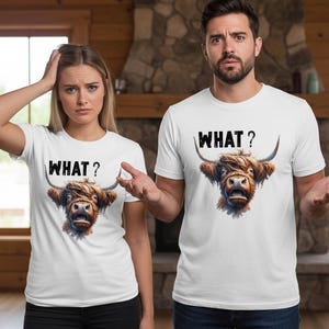 May include: Two people wearing white t-shirts featuring a Highland cow graphic and the word "WHAT?" in black. The cow graphic is brown and detailed. The background includes a stone fireplace and wooden paneling.