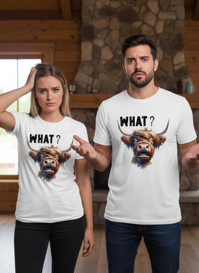 May include: Two people wearing white t-shirts featuring a Highland cow graphic and the word "WHAT?" in black. The cow graphic is brown and detailed. The background includes a stone fireplace and wooden paneling.
