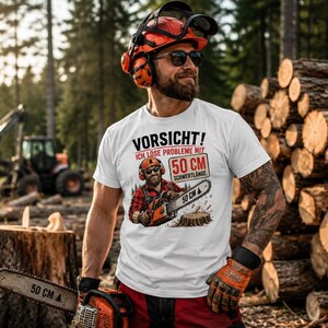 Puede incluir: Una camiseta blanca con el gráfico de un leñador con casco y protección auditiva sosteniendo una motosierra. El texto dice VORSICHT! ICH LÖSE PROBLEME MIT 50 CM SCHWERTLÄNGE. La hoja de la motosierra tiene impresa la medida de 50 CM.