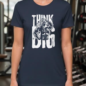 Könnte beinhalten: Marineblaues T-Shirt mit weißem Aufdruck. Der Aufdruck zeigt die Worte "THINK BIG" mit einer muskulösen Figur. Das Shirt ist aus weichem Material und eignet sich für das Fitnessstudio oder den Alltag.