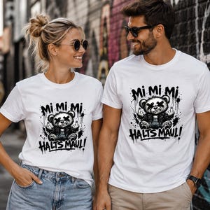 Könnte beinhalten: Ein weißes Kurzarm-T-Shirt mit einem schwarzen Grafikdruck eines Teddybären mit X-förmigen Augen, der eine Jacke trägt und den Mittelfinger zeigt. Der Text "Mi Mi Mi" steht über dem Bären und "HALTS MAUL!" ist darunter aufgedruckt.