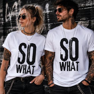 Könnte beinhalten: Weiße T-Shirts mit dem Text "SO WHAT" in fetten schwarzen Buchstaben. Die T-Shirts werden mit schwarzen Jeans und Accessoires getragen. Das Bild wird vor einer Graffiti-Wand aufgenommen.