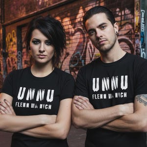 Könnte beinhalten: Zwei Personen tragen schwarze T-Shirts mit dem weißen Text "UNNU" und "FLENN MA NICH". Sie stehen vor einer mit Graffiti bedeckten Backsteinmauer. Die Person links hat dunkles Haar und die Person rechts hat kurzes dunkles Haar.