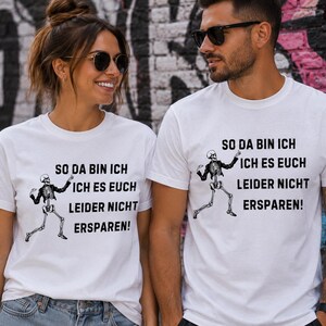 Könnte beinhalten: Ein weißes T-Shirt mit kurzen Ärmeln, das eine schwarze Grafik eines tanzenden Skeletts und den deutschen Text "SO DA BIN ICH ICH ES EUCH LEIDER NICHT ERSPAREN!" in einer fetten, schwarzen, serifenlosen Schrift auf der Vorderseite zeigt.