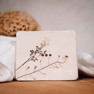 Könnte beinhalten: Quadratische, cremefarbene Seifenschale mit drei Abflusslöchern und gepressten botanischen Details. Die Schale zeigt zarte, getrocknete Pflanzenstängel und Blüten und schafft eine natürliche, rustikale Ästhetik.