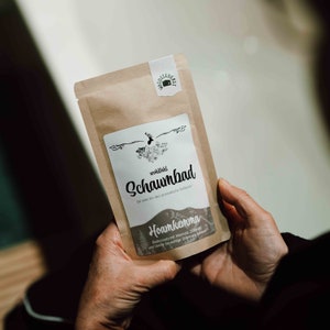 Könnte beinhalten: Eine braune Papiertüte mit einem weißen Etikett, auf dem "wohlfuhl Schaumbad" und "Hoamkemma" mit einer Berg- und Hirschillustration steht. Das Etikett sagt auch "Für zwei bis drei aromatische Vollbäder" und "Radicuzholz mit Melisse, Zirbe und Vanille für wohlige Stimmung".