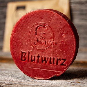 Könnte beinhalten: Eine runde, rote Blutwurst mit dem Wort "Blutwurst" geprägt auf der Vorderseite. Die Wurst hat ein kleines, geprägtes Bild eines menschlichen Gesichts in der Mitte.