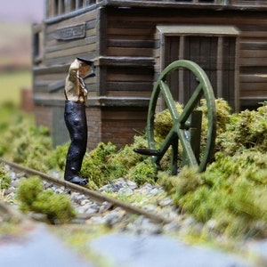 May include: A miniature model of a railway station with a small figure of a worker standing next to a green wheel. The station is made of wood and has a brown roof. The wheel is attached to a wooden structure and is partially obscured by green foliage.