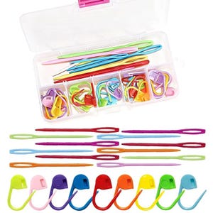 May include: A clear plastic storage box containing assorted colorful plastic yarn needles and stitch markers. Below the box, additional yarn needles in various colors are arranged horizontally, alongside a row of colorful plastic stitch markers.
