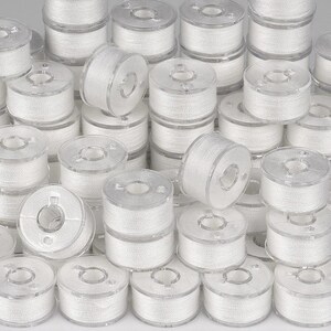 May include: A large collection of clear plastic sewing machine bobbins, each tightly wound with white thread. The bobbins are cylindrical with a central hole and small notches on the top and bottom rims, arranged in a dense, overlapping pile.