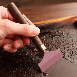 May include: A leather skiving tool featuring a dark brown wooden handle, a brass ferrule, and a wide, flat, maroon-colored metal blade. The tool is positioned against a dark, wet surface, highlighting the contrast between the wood and metal.