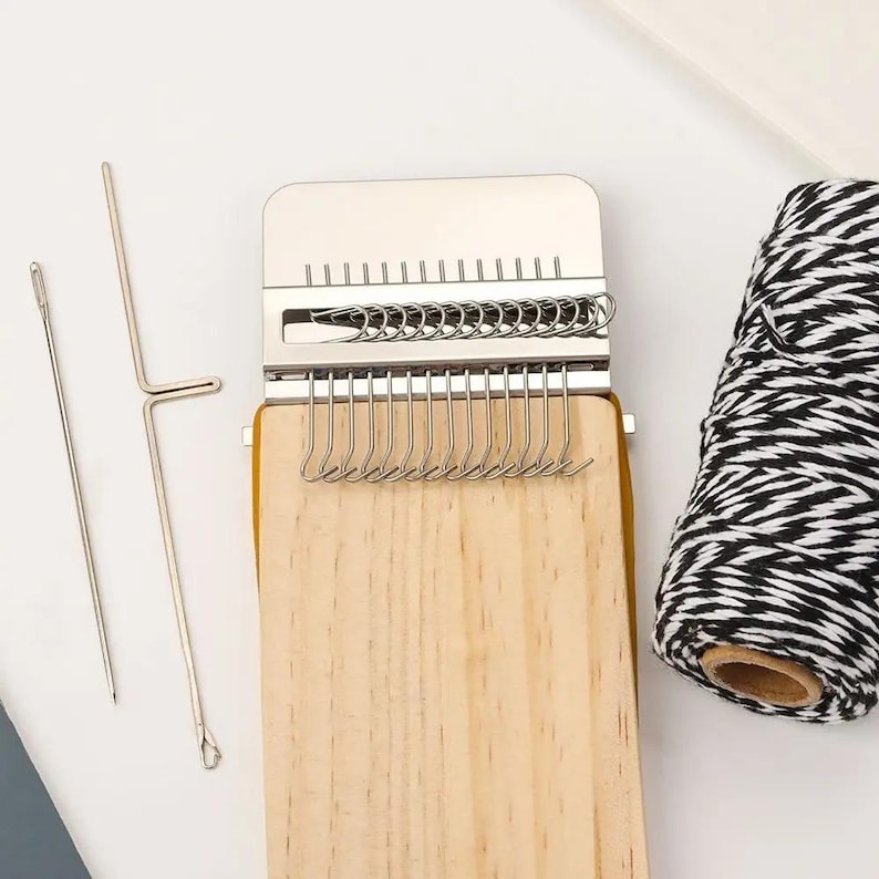 May include: A wooden weaving loom with a metal top and hooks, alongside two metal needles and a spool of black and white yarn. The loom has a light wood finish and is designed for crafting.