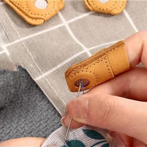 May include: Close-up of a tan leather finger protector for sewing. The finger protector has a reinforced tip with a small mesh circle and is being used to guide a needle through fabric. The image shows the finger protector in use.