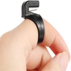 May include: A black plastic ring-style yarn cutter featuring a small, sharp metal blade integrated into the top section. The circular band is designed to fit around a finger, with the blade positioned to cut thread or yarn during crafting tasks.