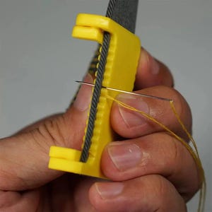 May include: A yellow needle threader tool with a gray cord threaded through it. A needle with yellow thread is being inserted through the cord. The tool is held in a person's hand.