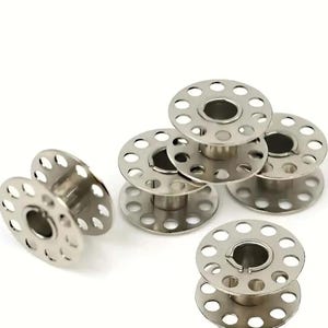 May include: A collection of silver metal sewing machine bobbins. Each bobbin has a circular design with multiple small holes around the perimeter and a central hole. The bobbins are stacked and arranged on a white background.